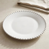 An empty white plate with a raised beaded rim rests on a beige surface beside a folded cloth.