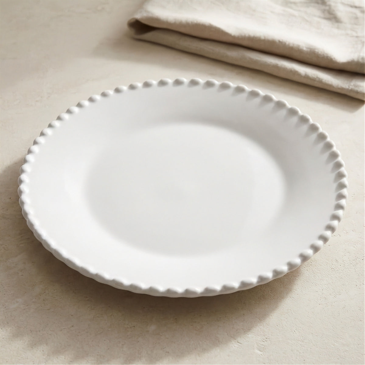 An empty white plate with a raised beaded rim rests on a beige surface beside a folded cloth.