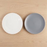 A white beaded-rim plate and a smooth gray round plate sit side by side on a light wooden surface.