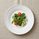 White plate with a beaded rim holds leafy greens, cherry tomato halves, and cucumber slices on beige textured fabric.
