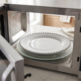 White plate with a beaded rim rests on the glass turntable inside an open microwave on a kitchen counter.