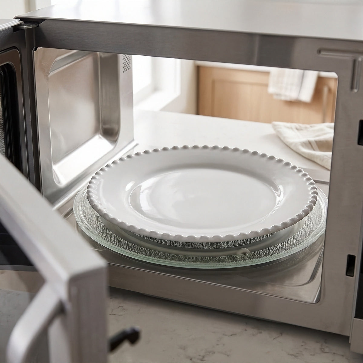 White plate with a beaded rim rests on the glass turntable inside an open microwave on a kitchen counter.