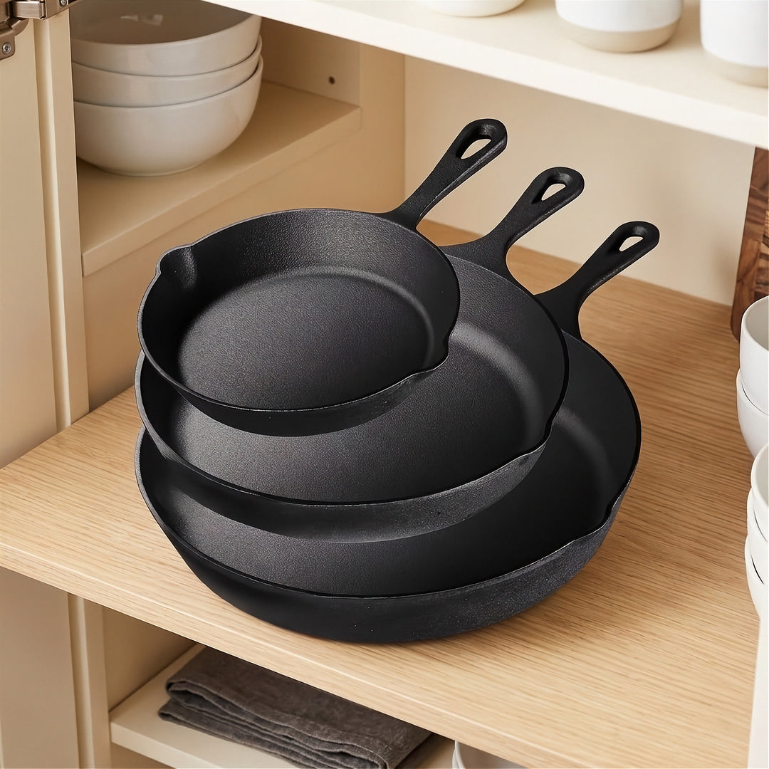 Three black cast iron skillets nest on a wooden cabinet shelf beside stacked white bowls.