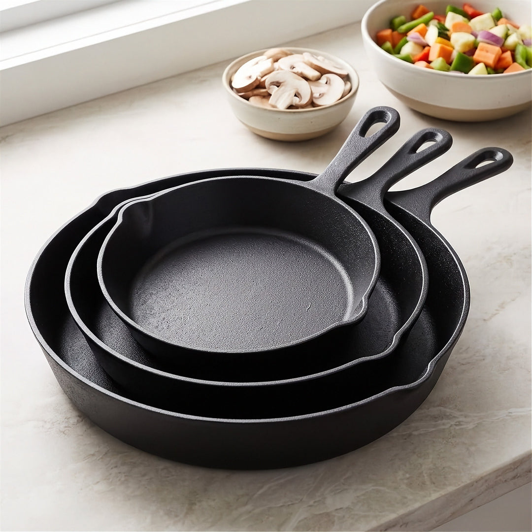 Three black cast iron skillets nest on a pale countertop beside bowls of sliced mushrooms and chopped vegetables.