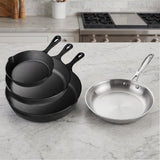 Three nested black cast iron skillets stand beside a silver frying pan on a pale kitchen countertop.