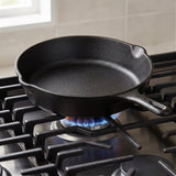 Black cast iron skillet sits over a blue gas flame on stove grates, with a light tiled backsplash behind it.