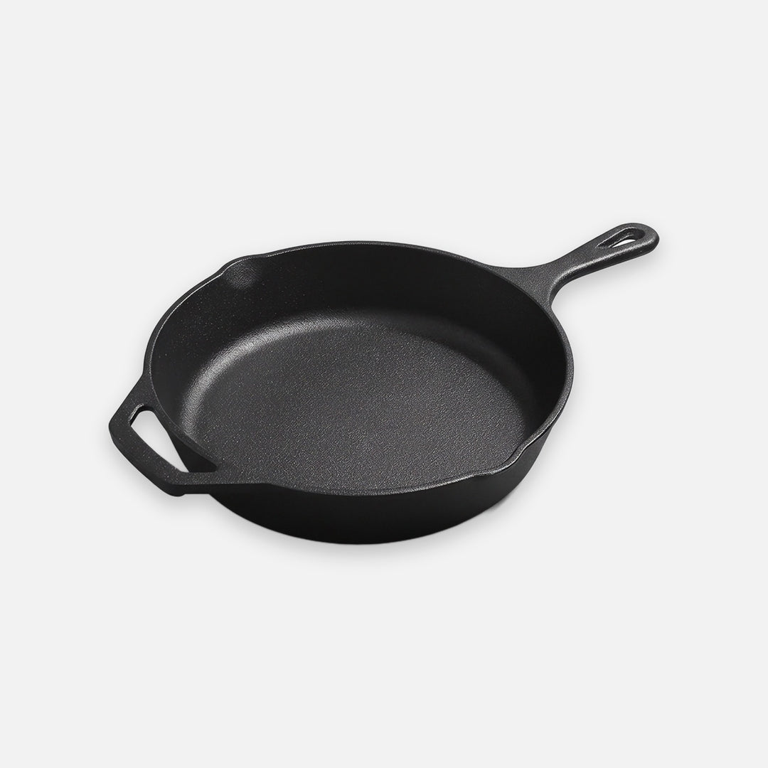 A black cast iron skillet with a long handle, helper handle, and pouring spouts sits against a light background.