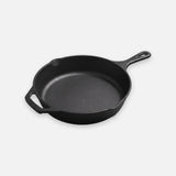 A black cast iron skillet with a long handle, helper handle, and pouring spouts sits against a light background.