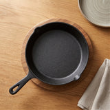 A black cast iron skillet rests on a round wooden board beside a beige napkin and gray plate.