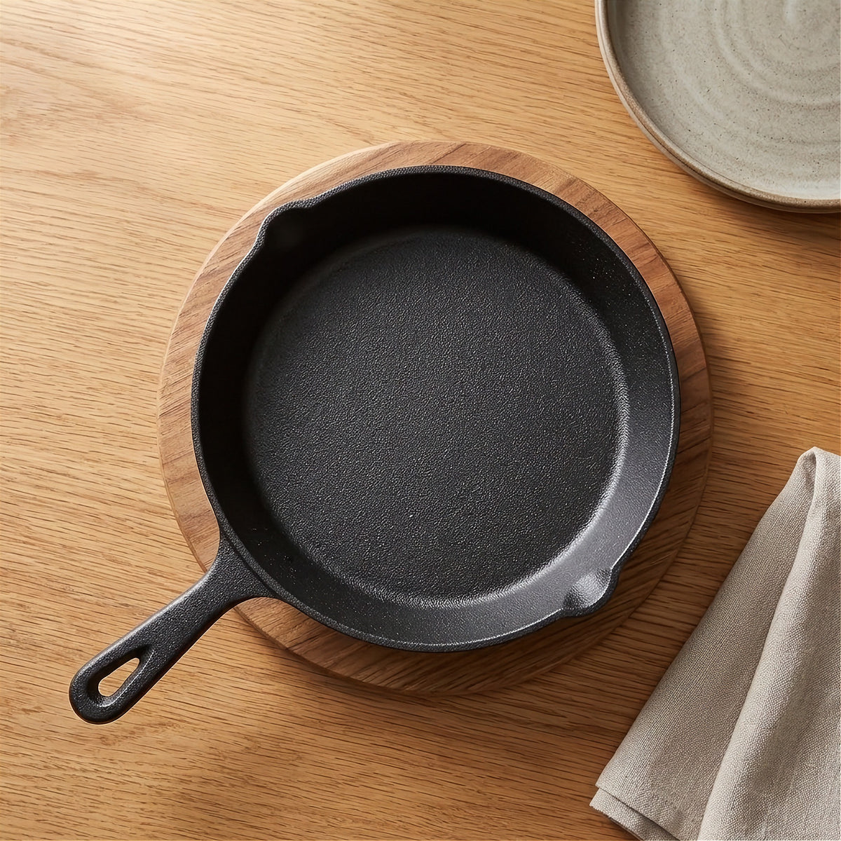 A black cast iron skillet rests on a round wooden board beside a beige napkin and gray plate.