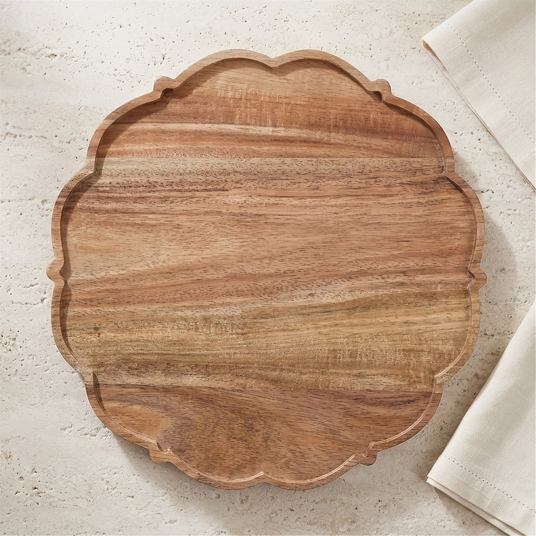 An oval wooden serving board with a scalloped edge and visible wood grain, photographed from above on a light textured surface with a folded white cloth partially visible at the right edge.