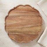 An oval wooden serving board with a scalloped edge and visible wood grain, photographed from above on a light textured surface with a folded white cloth partially visible at the right edge.