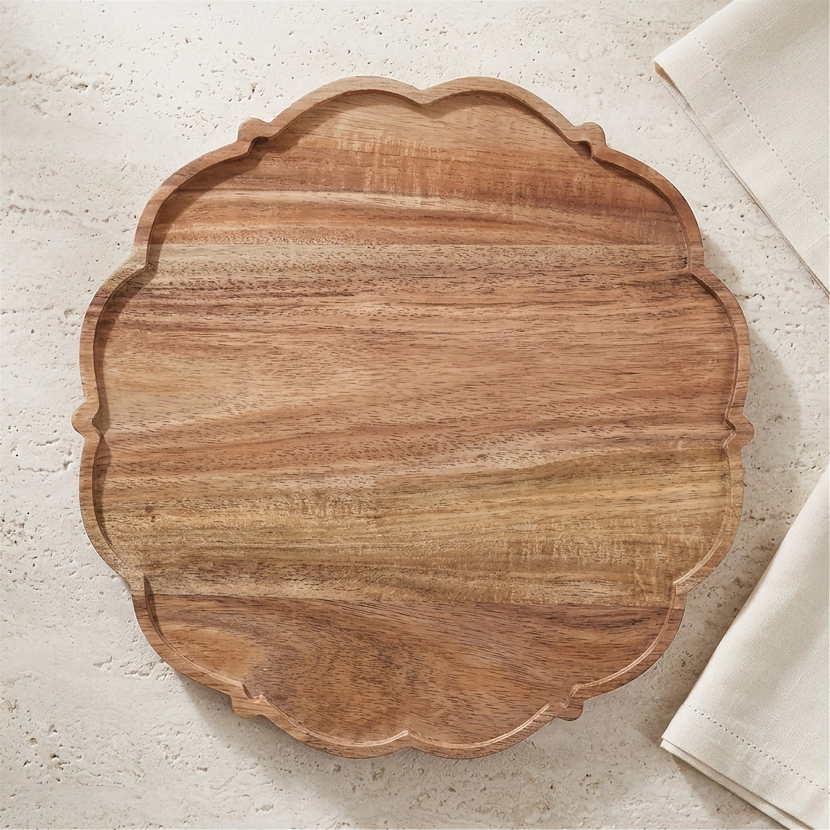 An oval wooden serving board with a scalloped edge and visible wood grain, photographed from above on a light textured surface with a folded white cloth partially visible at the right edge.