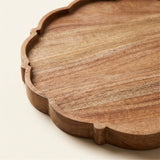 A close-up of a scalloped wooden serving tray with a warm medium-brown wood grain finish, set against a clean off-white background.