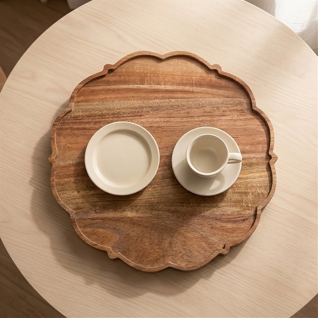 An oval wooden serving tray with scalloped edges, styled on a round light wood table with a cream-colored ceramic plate and cup on top.