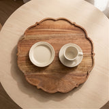 An oval wooden serving tray with scalloped edges, styled on a round light wood table with a cream-colored ceramic plate and cup on top.