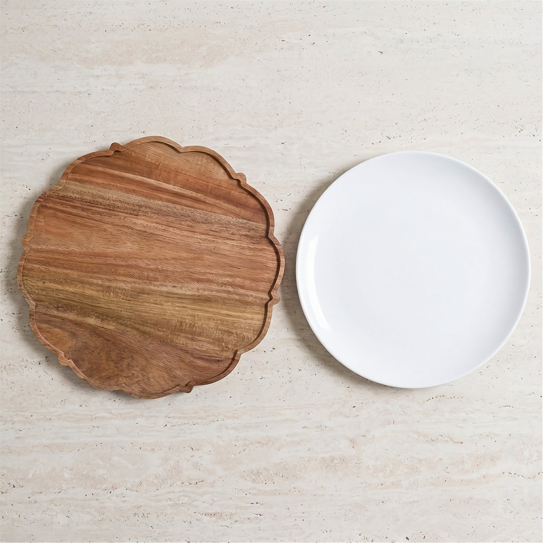 A scalloped wooden serving board with a warm brown wood grain finish and a smooth white ceramic plate, displayed on a light beige stone surface.