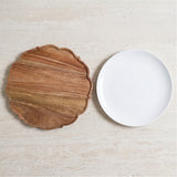 A scalloped wooden serving board with a warm brown wood grain finish and a smooth white ceramic plate, displayed on a light beige stone surface.