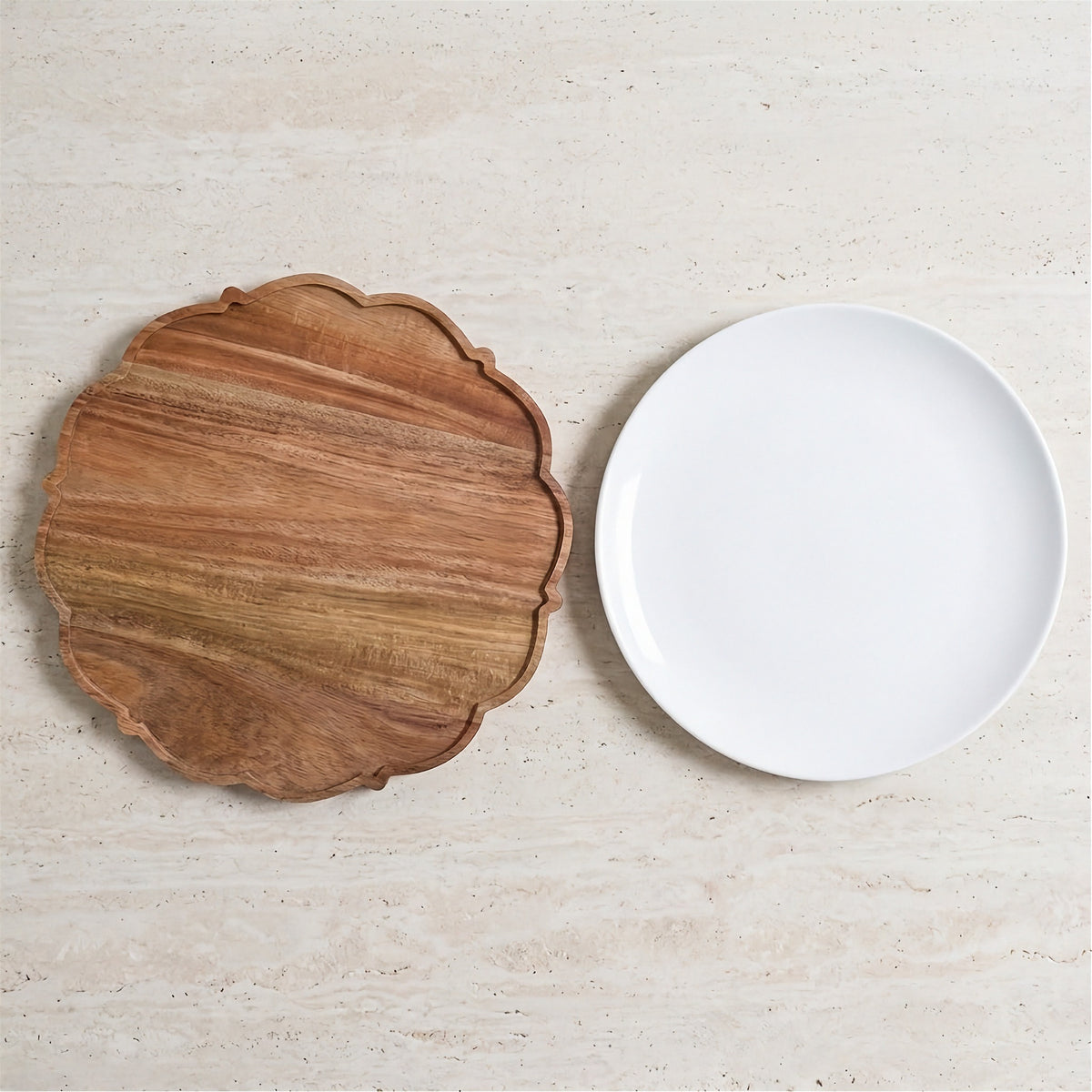 A scalloped wooden serving board with a warm brown wood grain finish and a smooth white ceramic plate, displayed on a light beige stone surface.