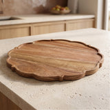 A scalloped-edge round wooden serving board with visible wood grain, placed on a light beige stone countertop in a kitchen setting.