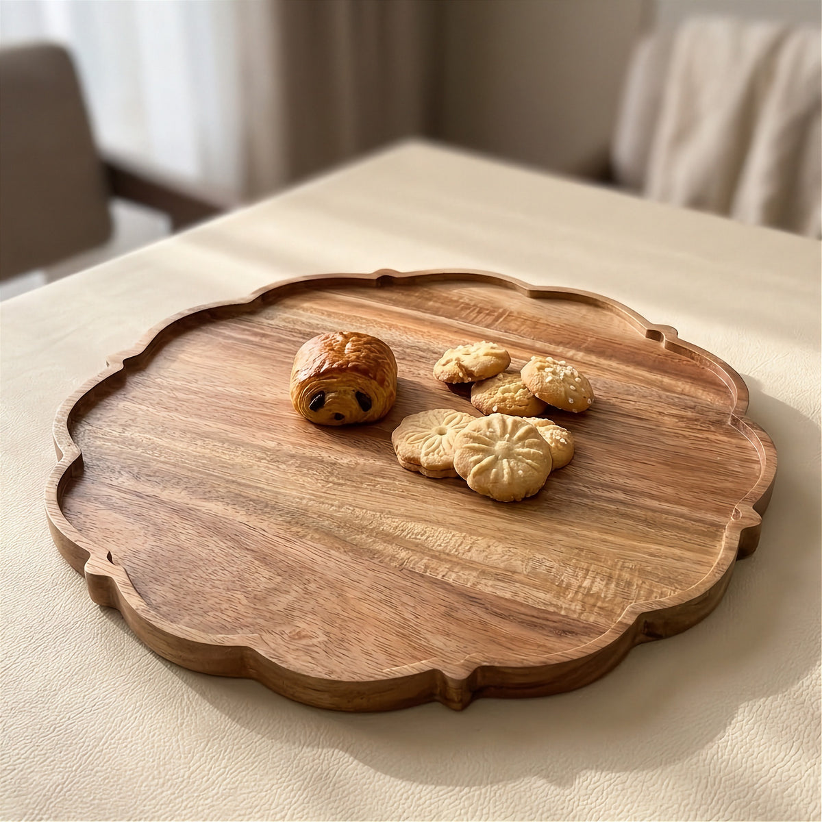 A scalloped wooden serving tray with a warm medium-brown finish, styled with a small pastry and cookies on a beige tablecloth in a softly blurred indoor setting.