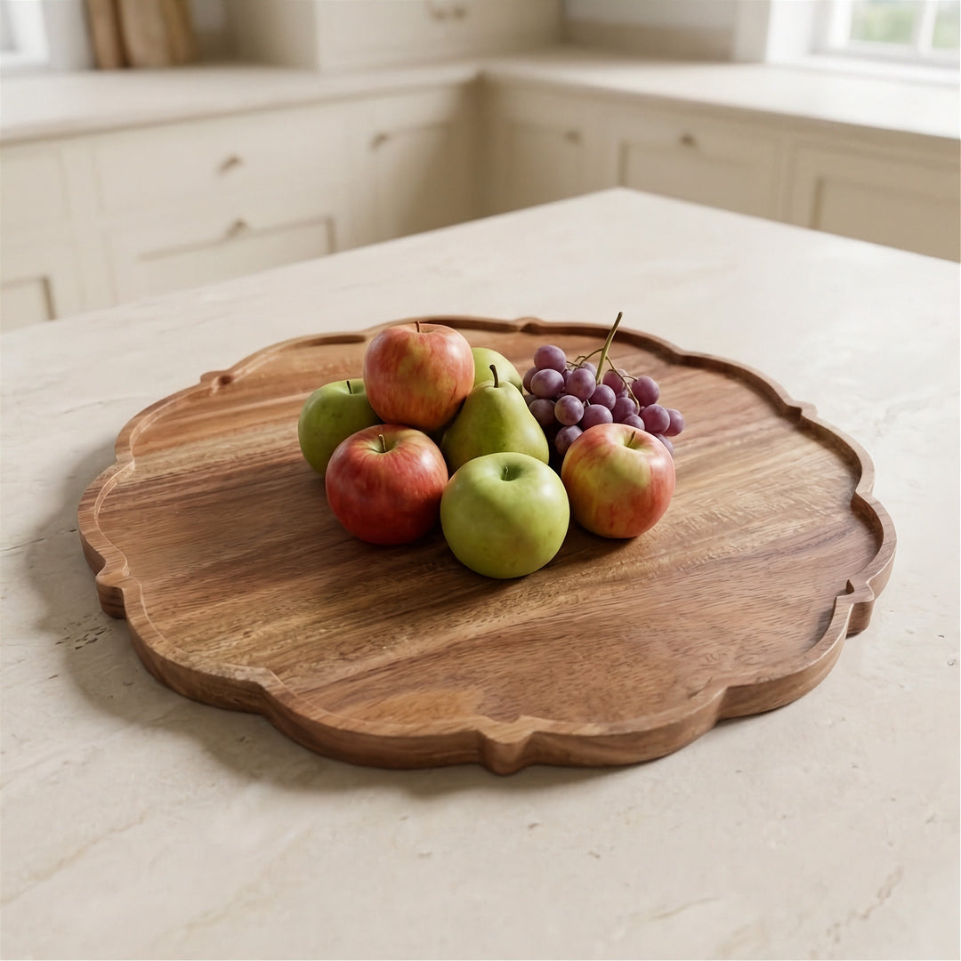 A scalloped round wooden serving board on a light stone countertop, styled with fresh apples, pears, and a bunch of purple grapes in a bright kitchen setting.