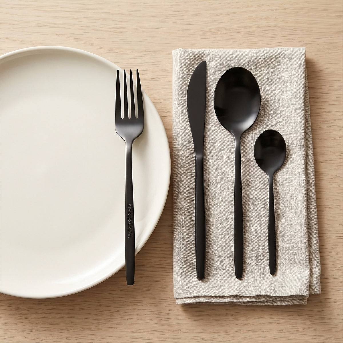 Black flatware set including a fork, knife, spoon, and teaspoon arranged on a beige linen napkin next to a white plate on a light wood table.