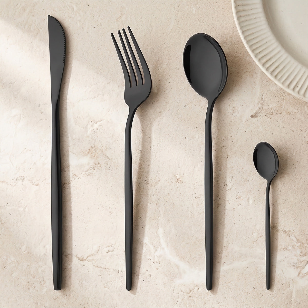 Four-piece matte black cutlery set including a knife, fork, dinner spoon, and teaspoon, arranged on a light beige stone surface next to a white plate edge.