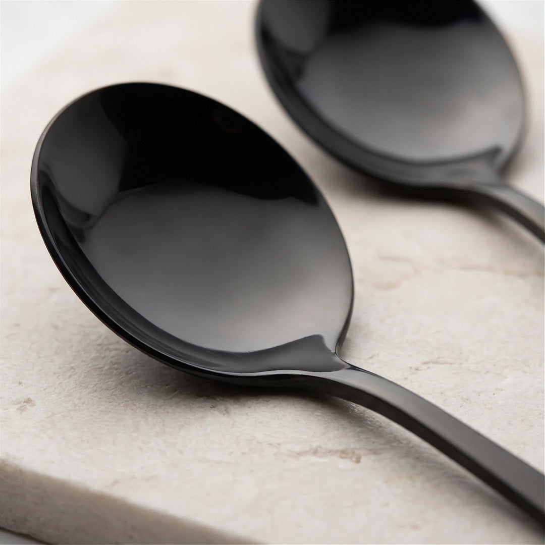 Two glossy black metal spoons resting on a light beige stone surface, with one spoon in sharp focus in the foreground and the other softly blurred in the background.
