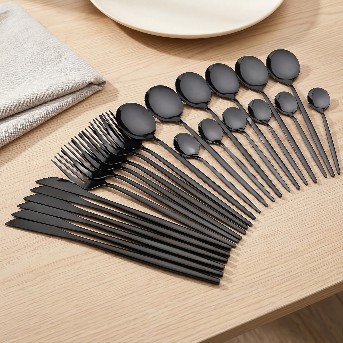 A set of matte black flatware, including spoons, forks, and knives, arranged in a fan shape on a light wood tabletop next to a folded beige napkin and a white plate.