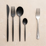 Five-piece flatware set arranged on a light beige stone surface, including a black knife, black plastic fork, black spoon, small black spoon, and a silver metal fork.