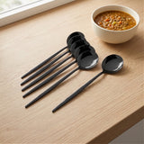 Seven black metal spoons arranged on a light wood tabletop next to a white bowl of soup, with a window visible in the background.