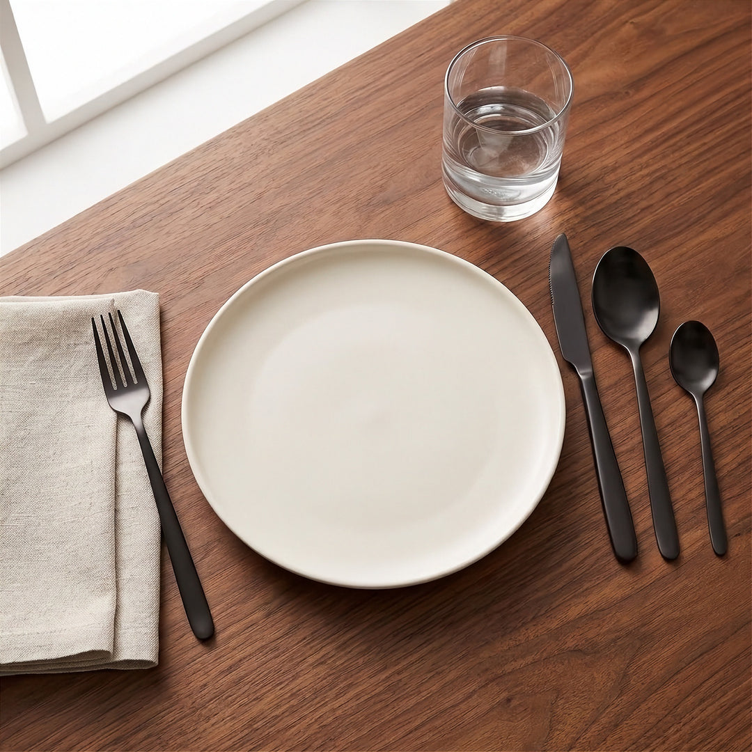 A minimalist beige ceramic plate set on a dark wood table with matte black cutlery, a folded beige napkin, and a clear drinking glass.