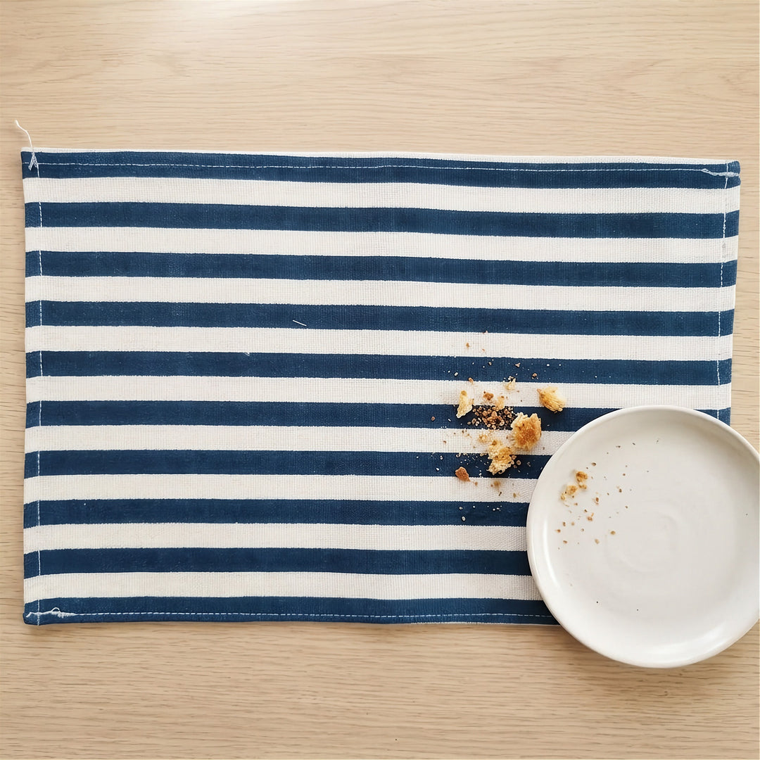 A rectangular fabric placemat with horizontal navy blue and off-white stripes, styled on a light wood tabletop next to a white ceramic plate with crumbs.
