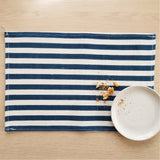 A rectangular fabric placemat with horizontal navy blue and off-white stripes, styled on a light wood tabletop next to a white ceramic plate with crumbs.