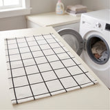 A rectangular white kitchen towel with a black grid pattern, laid flat on a light beige countertop in a bright laundry room next to a washing machine.