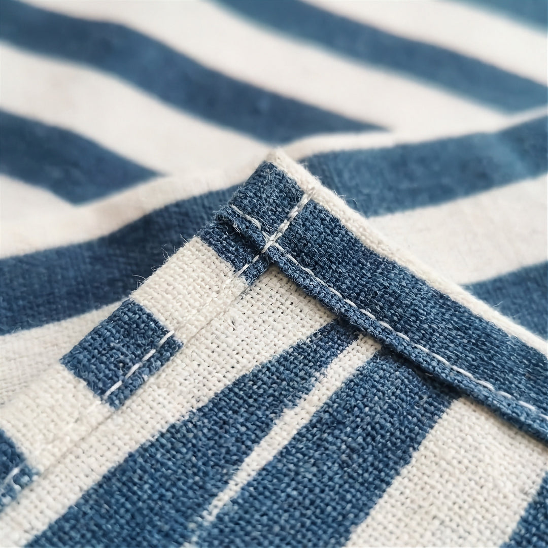 Close-up of a woven textile with a blue and off-white striped pattern, showing the fabric texture and a folded corner with visible stitching.