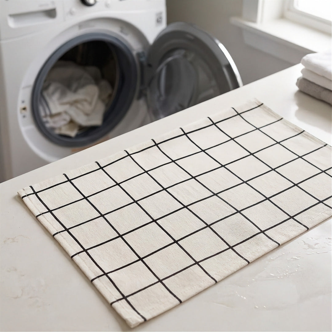 A cream-colored kitchen towel with a black grid pattern laid out on a white countertop in a laundry room, with a washing machine and folded towels visible in the background.