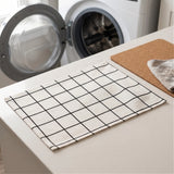 A white kitchen towel with a black grid pattern laid out on a light countertop, with a washing machine blurred in the background and a cork mat beside it.