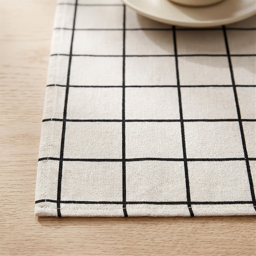 A close-up of a woven placemat with a black grid pattern on a cream background, laid on a light wood table with a partial view of a ceramic cup and saucer in the top right corner.