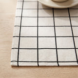 A close-up of a woven placemat with a black grid pattern on a cream background, laid on a light wood table with a partial view of a ceramic cup and saucer in the top right corner.