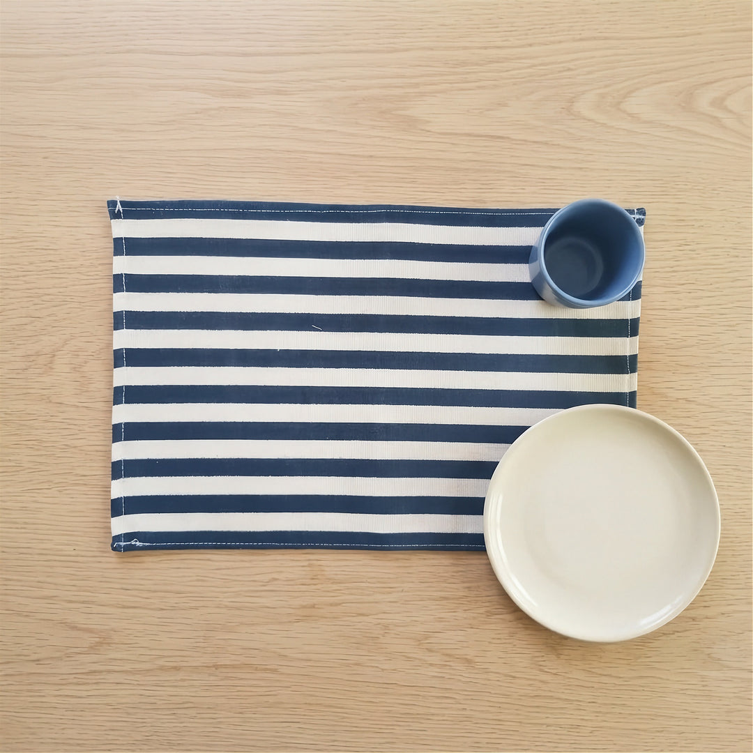 A navy blue and white striped fabric placemat on a light wood tabletop, styled with a small blue cup and a white plate.