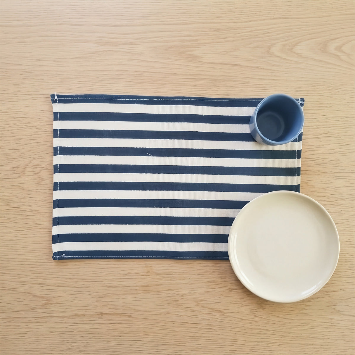 A navy blue and white striped fabric placemat on a light wood tabletop, styled with a small blue cup and a white plate.
