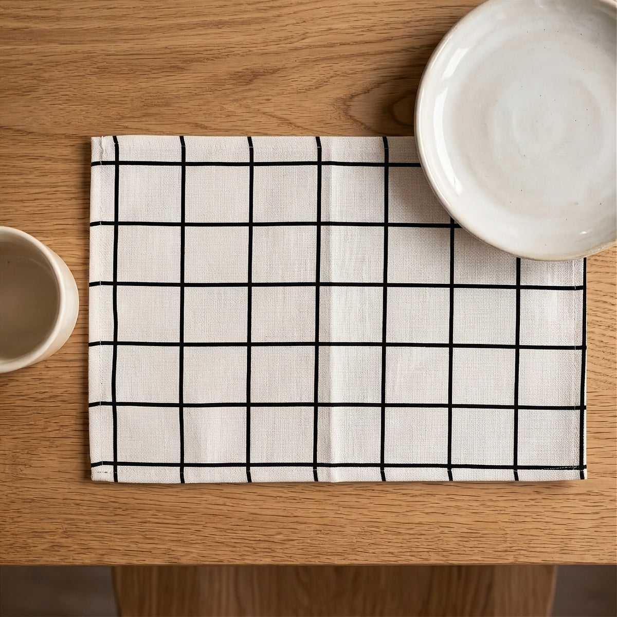 A square white linen placemat with a black grid pattern, styled on a wooden table with ceramic dishes partially visible in the corners.