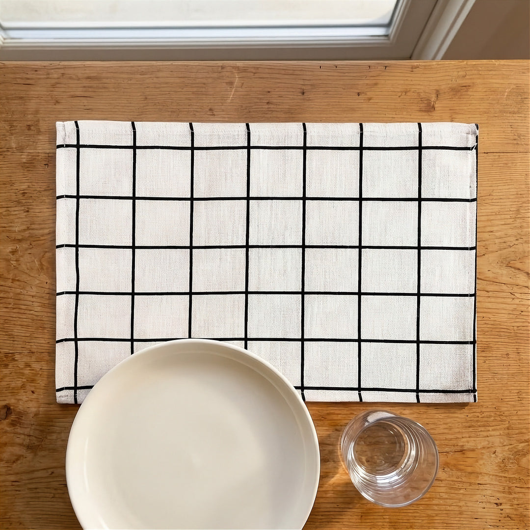 A rectangular white linen placemat with a black grid pattern, laid on a wooden table with a white ceramic plate and a clear glass partially visible in the foreground.