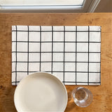 A rectangular white linen placemat with a black grid pattern, laid on a wooden table with a white ceramic plate and a clear glass partially visible in the foreground.