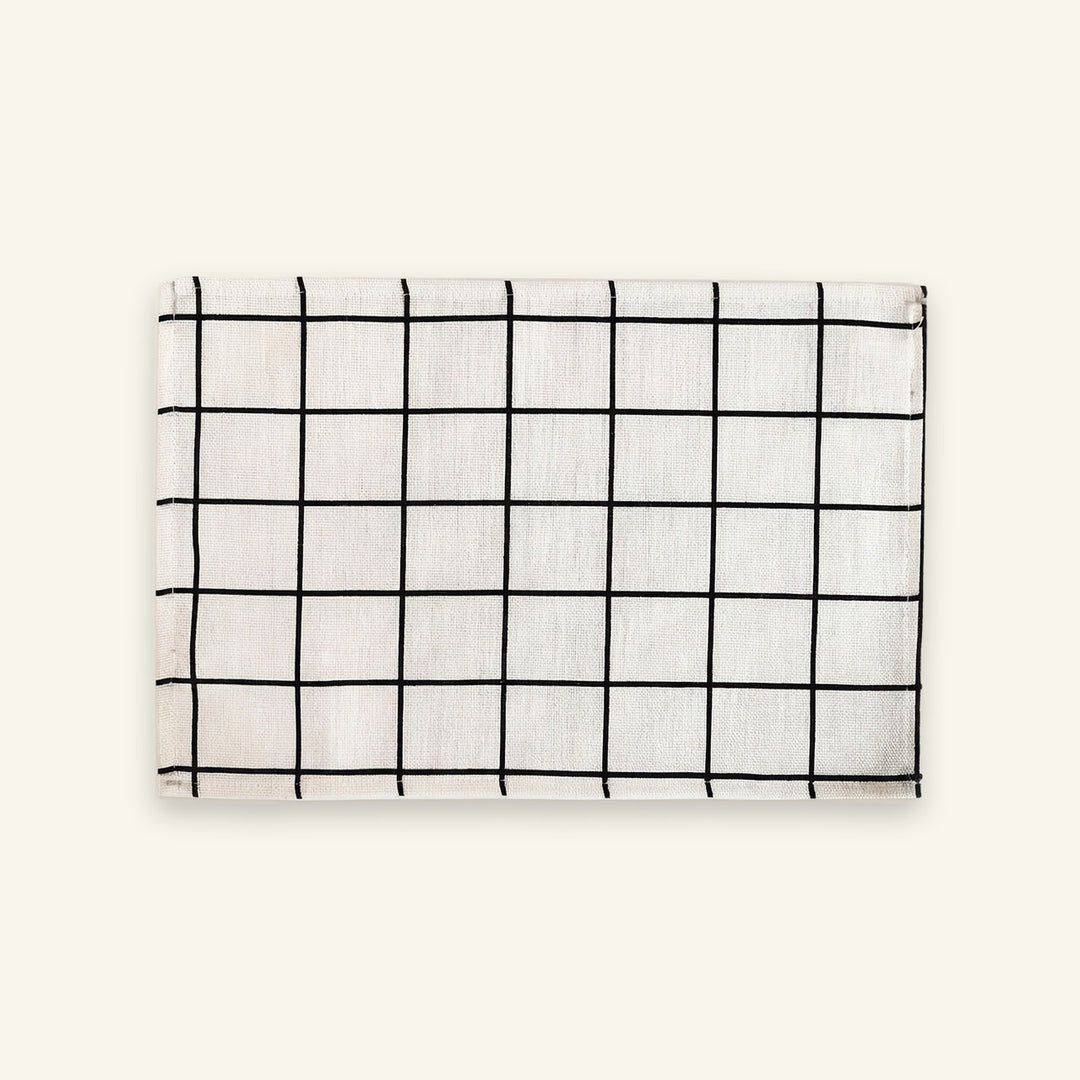 A rectangular white fabric item, likely a napkin or small towel, featuring a black grid pattern, displayed flat against a solid off-white background.