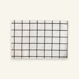 A rectangular white fabric item, likely a napkin or small towel, featuring a black grid pattern, displayed flat against a solid off-white background.