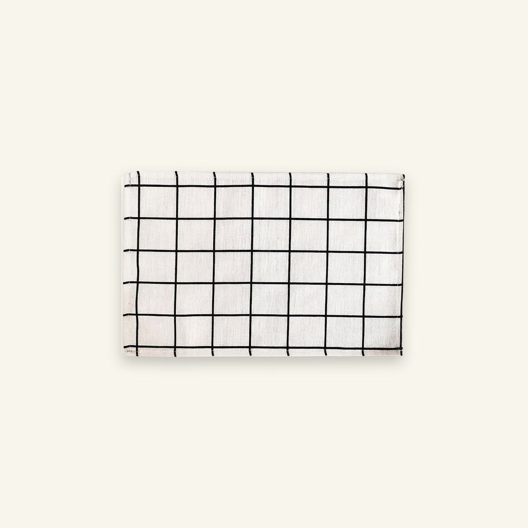 A rectangular white fabric item, likely a napkin or small towel, featuring a black grid pattern, displayed against a solid off-white background.