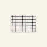 A rectangular white fabric item, likely a napkin or small towel, featuring a black grid pattern, displayed against a solid off-white background.